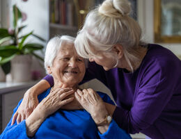 An older woman hugs a seated older woman.