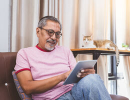 A seated man uses a tablet as a cat watches from a nearby shelf.