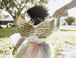A little girl in a glittery dress with gold wings walks down a street.