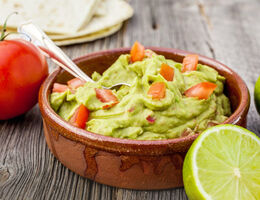 A bowl of guacamole surrounded by limes and a tomato.