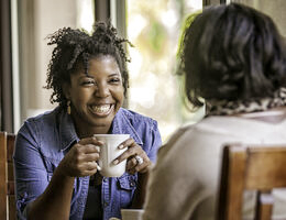 A woman holding a mug sits in a coffee shop with a friend.