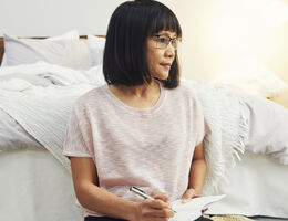 A pensive woman leans against the foot of a bed.