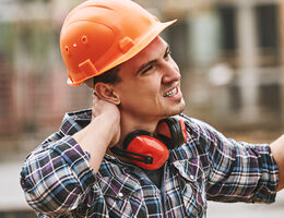 A man in a hard hat, wearing his earmuffs lowered, rubs his neck.