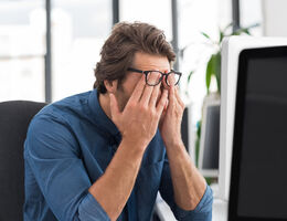 A man at an office desk leans forward and rubs his eyes.