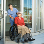  A woman in scrubs pushes a patient in a wheelchair out the door of a hospital.
