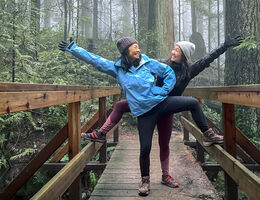 Two women in cool-weather hiking clothes pose on a bridge in the woods.