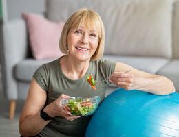 A woman leans on an exercise ball as she eats a salad.