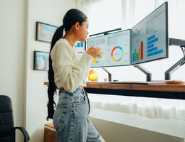 A woman at a standing desk holds a glass and looks at her monitors.