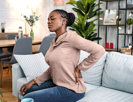 A woman sits on a couch, rubbing her sore back