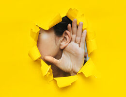 A hand cupping an ear, seen through a torn hole in a yellow paper wall.