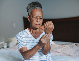 A woman sits on a bed and holds her raised wrist.