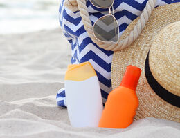 A pair of sunscreen bottles in the sand beside a beach bag and a straw hat.