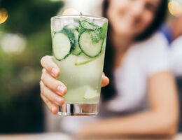 A woman holds a glass of clear liquid with mint leaves and sliced pear and cucumber.