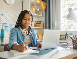 An adolescent writes in a notebook while sitting in front of an open laptop.