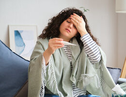 A woman on a couch holds a hand to her forehead as she looks at a thermometer.